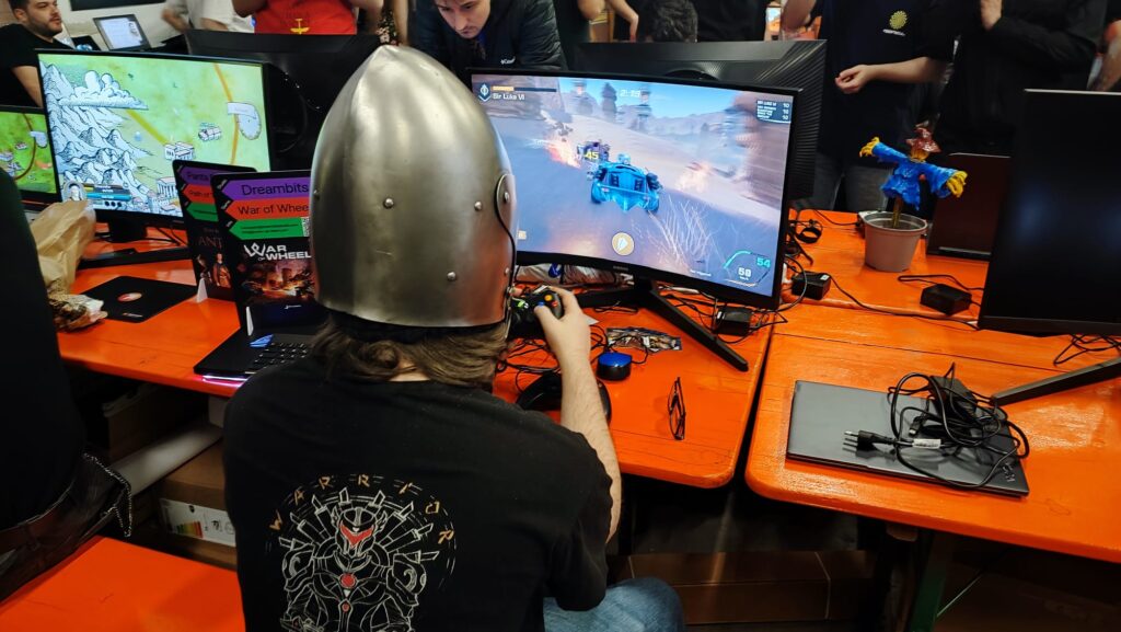 Giocatore con elmo medievale e controller in mano partecipa a un evento di videogiochi a postazione con PC.